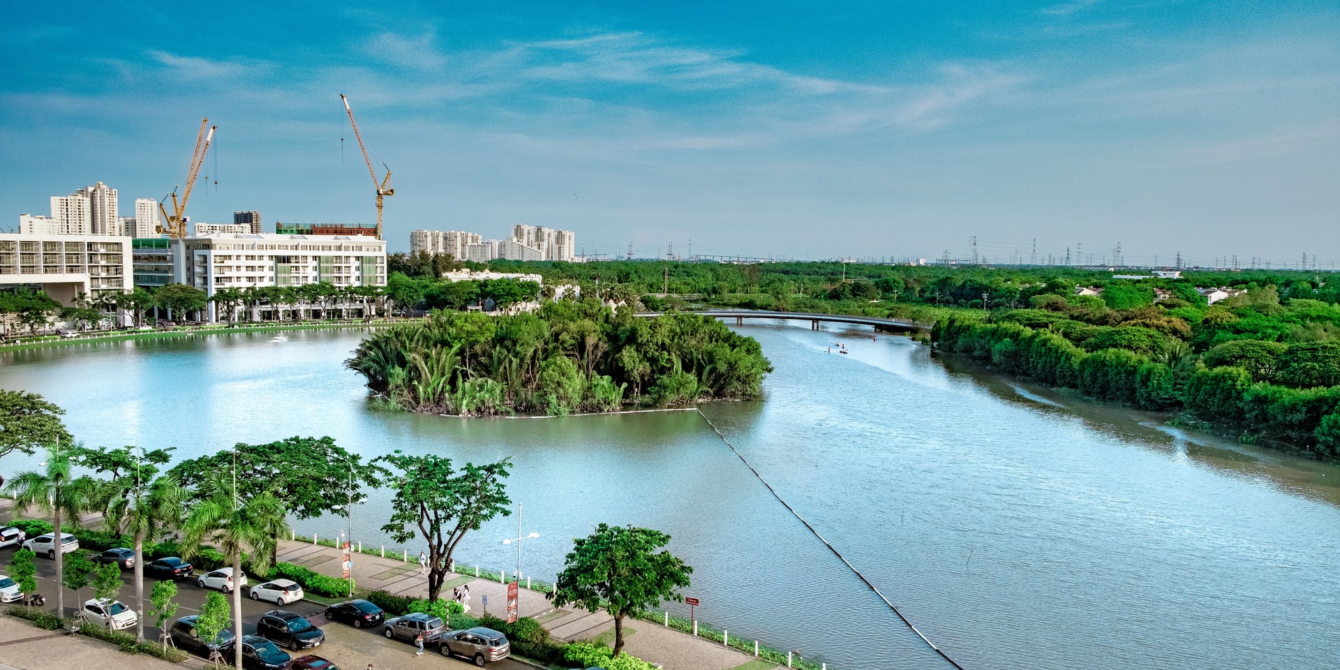 A scenic view of a river with lush green trees, surrounding buildings under construction, and vehicles parked on a road beside the water.