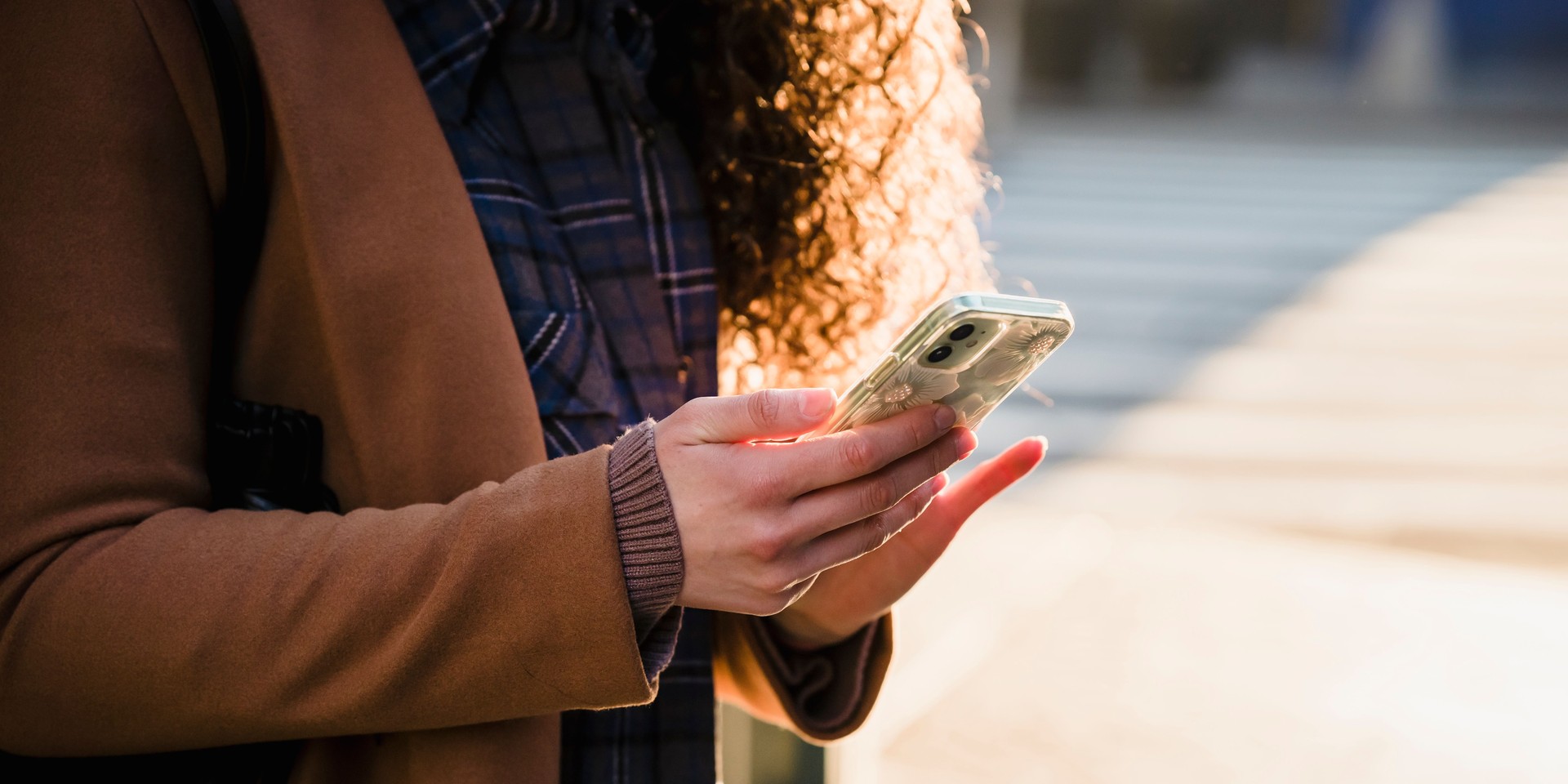 A person in a brown coat holds a smartphone with a floral case, standing outdoors in sunlight.