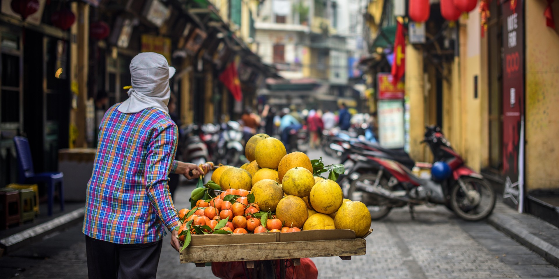 A person in a plaid shirt and covered head pushes a cart with pomelos and oranges through a narrow street, surrounded by motorcycles and buildings.