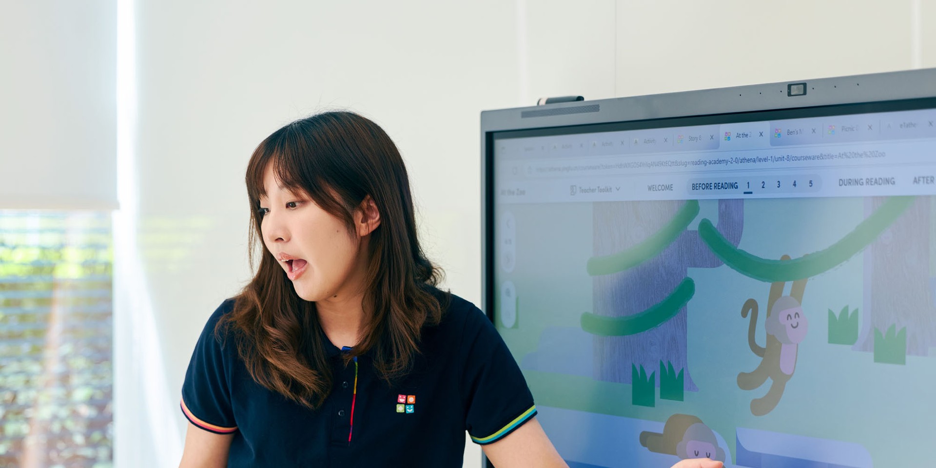 A woman in a navy shirt points at a digital screen displaying a playful monkey illustration, while explaining something.
