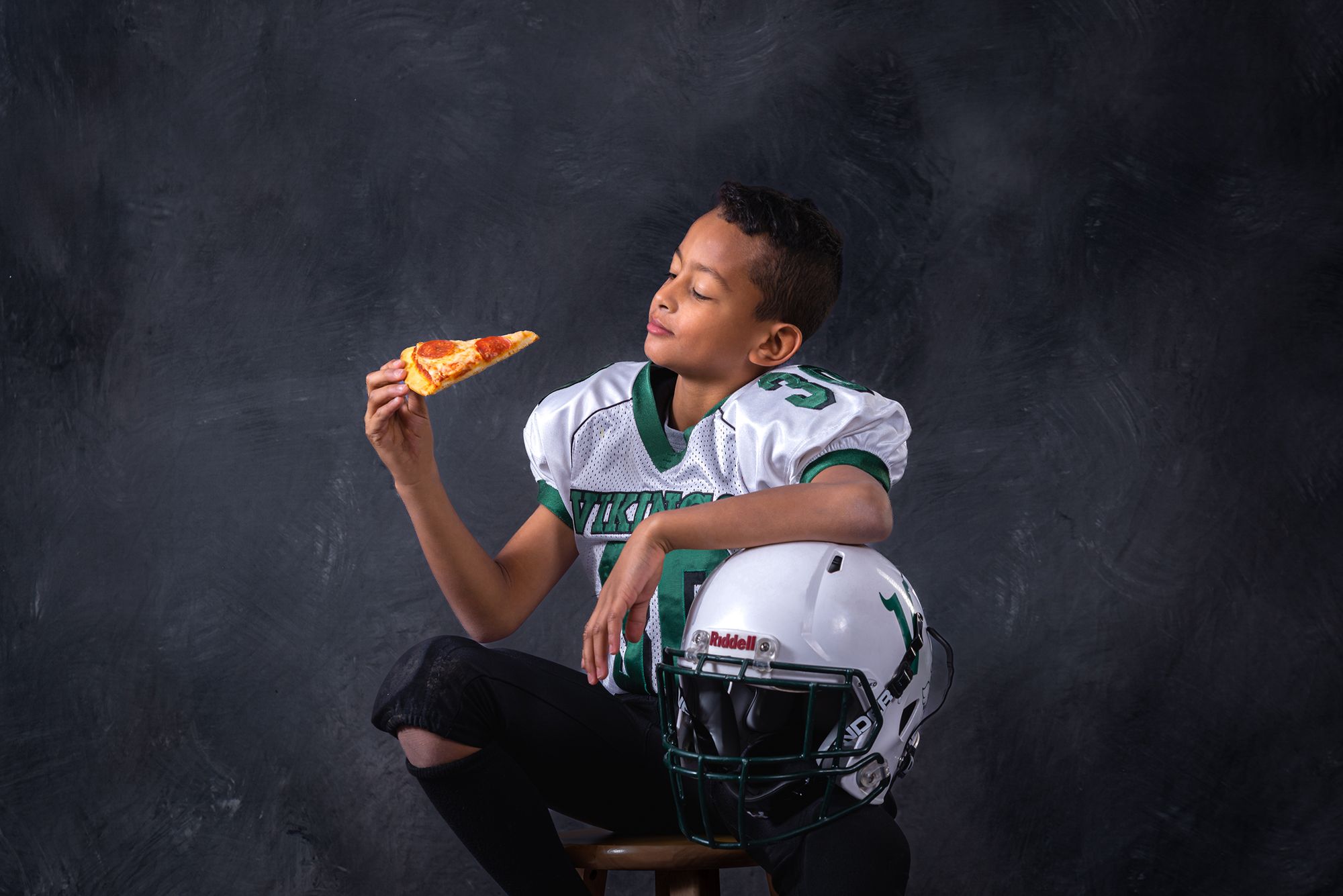 boy in football gear sitting on a chair, ready to bite into a pizza slice