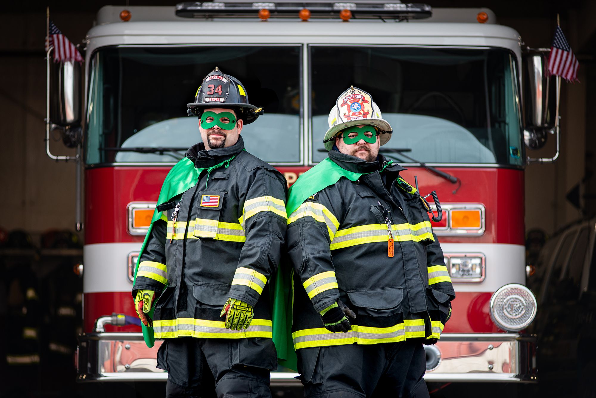 two firemen in firefighting gear plus green Hoagie Hero capes posing in front of firetruck