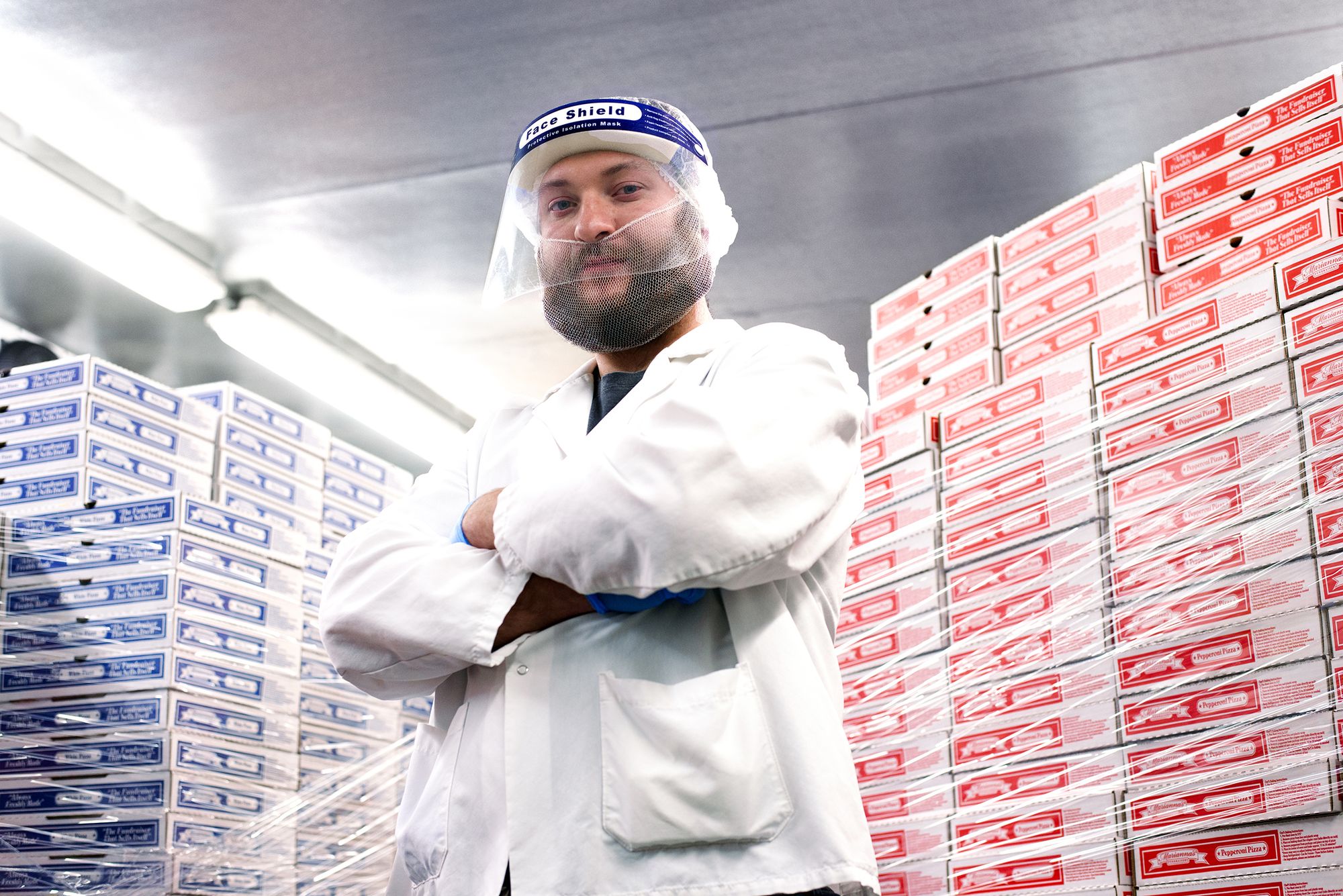 marianna's employee wearing white coat, face shield, beard and hair net posing in front of stacks of pizza boxes