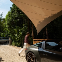 Beige schaduwdoek boven een oprit met een geparkeerde sportwagen en een voorbijlopende persoon.
