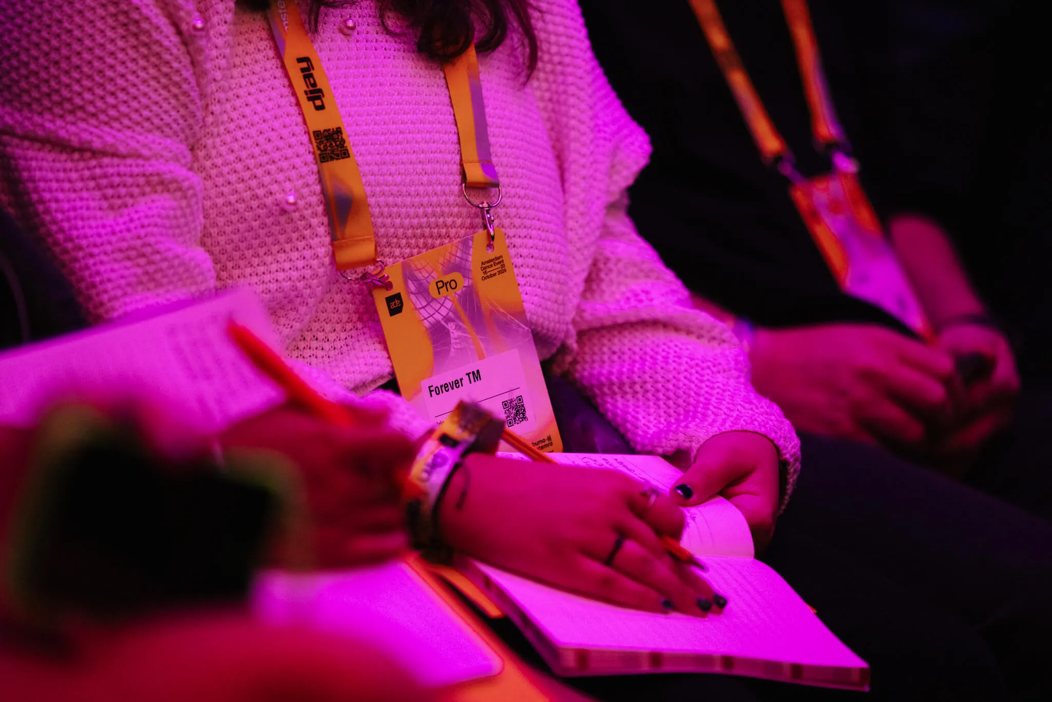 Person taking notes in a notebook at ADE panel by MassiveMusic, wearing a white sweater and yellow lanyard, under purple lighting.