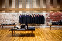Interior of a store. Wooden benches are placed on top of a wooden floor showcasing a variety of products including shoes and bags. Black suit jackets are hung neatly along a long black hanger in front of a brick wall. Above the brick wall, wooden Fortina panels are laid vertically with one large strip of wood running horizontally across them.