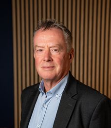 A man in a blue polka dot shirt stands in a modern office with wooden wall panels and plants in the background.