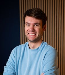 Smiling person with short hair wearing a brown sweater, standing in front of a wooden wall with small potted plants.