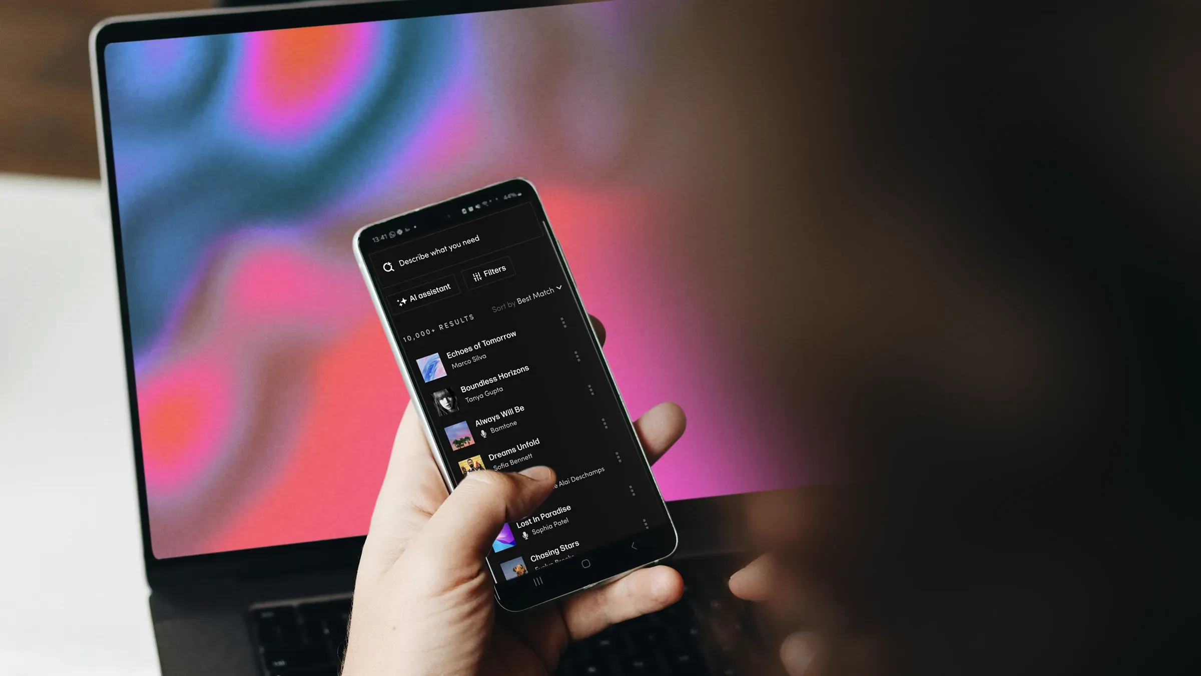 Hand holding a phone running a music search app in front of a laptop, showing music built into digital platforms