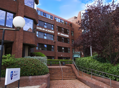 Brick building of Dublin Academy of Education with large windows, surrounded by greenery, and a sign in front.