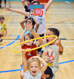 Children enthusiastically playing with a yellow hoop inside a gymnasium, with arms raised and smiling faces.
