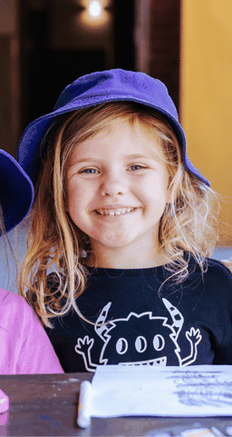 A smiling child with long blonde hair wearing a purple hat and a black shirt with a monster design, sitting at a table.