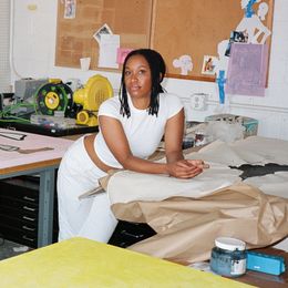 Tschabalala Self, wearing white, leaning on a table in her New Haven studio