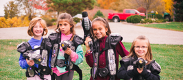 Four children wearing laser tag gear, smiling and holding phasers, standing on grass with autumn trees and cars in the background.