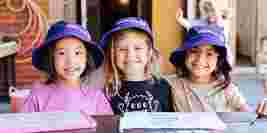 Three young children wearing OSHClub hats, smiling at a table with drawing pads, against a brick wall backdrop.