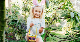 Girl wearing bunny ears, kneeling in a garden, holding an Easter egg and a basket with eggs, surrounded by green foliage.