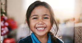 Smiling child with dark hair, wearing a blue shirt against a softly lit, blurred background.
