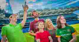 A group of people stands on a sports field, one person pointing upwards, with seating and a scoreboard visible in the background.