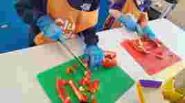 Children wearing aprons and gloves slice red bell peppers on colorful cutting boards in a cooking class.