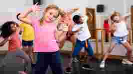 Children in colorful clothes dancing energetically in a studio, with a focus on a girl in a pink shirt smiling at the camera.
