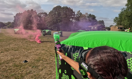 Person playing gel blaster game outdoors, aiming at opponents hiding behind inflatable barriers, with colored smoke in the background.