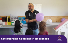 Man holding a purple balloon speaks to three children in a classroom, next to a whiteboard and colorful books. Text: "Safeguarding Spotlight: Meet Richard."