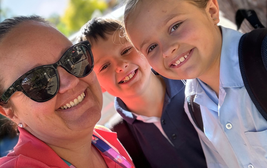 A woman wearing sunglasses smiles with two children, both in school uniforms, outdoors on a sunny day.