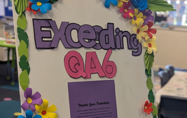 Decorative board with "Exceeding QA6" in colorful letters and paper flowers, thanking families for support.