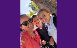 Smiling adult and two children pose for a selfie outdoors on a sunny day, with a large tree in the background.