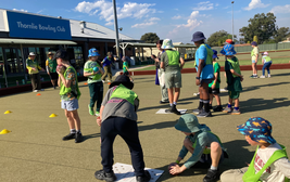 Children playing lawn bowls at Thornlie Bowling Club on a sunny day, wearing colorful hats and green vests.