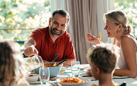 Family enjoying a meal together, with a man pouring water and smiling, while three others sit around a table with pasta dishes.