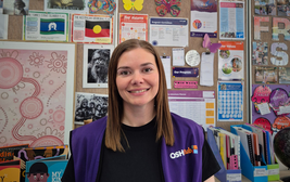 A person smiling in front of a colorful bulletin board filled with posters and information, wearing a purple vest with "OSHClub" logo.