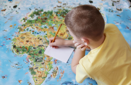 A child in a yellow shirt writes on a piece of paper while leaning over a colorful world map with animals and landmarks.