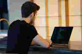 A man in a black shirt sits and codes on a laptop screen