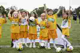A group of young children in yellow shirts and orange shorts celebrate on a soccer field, holding soccer balls and raising their arms in excitement.