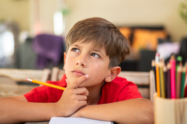 A young boy in a red shirt sits at a table, deep in thought, holding a pencil to his chin, with colorful pencils in a holder nearby.
