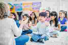A group of young children sitting cross-legged on a classroom floor, facing a teacher who is clapping. Bright posters decorate the walls.
