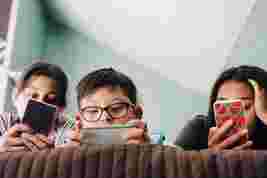 Three children sitting closely, each absorbed in a smartphone. They are indoors, focused, with a patterned wall in the background.