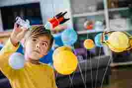 A child in a yellow shirt plays with a model solar system, holding a toy astronaut and red rocket while looking up intently.