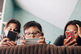 Three kids focused on their smartphones, sitting together on a sofa with a patterned teal wall in the background.