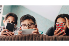 Three children lying on a couch, focused on their smartphones and a tablet, with a colorful, patterned background behind them.