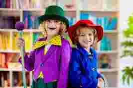 Two children in colorful costumes, one in purple with a green hat and cane, and the other in blue with a red hat, smiling in a library setting.