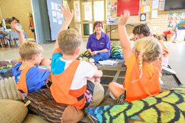 Children sitting with a teacher in a classroom, some raising their hands. The room has colorful decorations and educational materials on the walls.