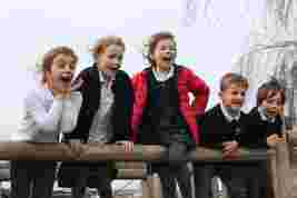 Five children leaning over a wooden railing, smiling and cheering on a windy day.