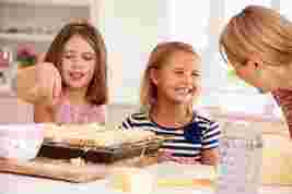 Two young girls and a woman in a bright kitchen, smiling and preparing cheesy bread on a tray.