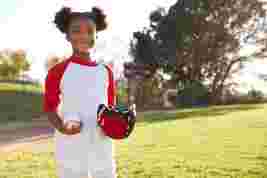 A smiling child in a baseball field, wearing a red and white shirt, holding a baseball and glove, standing on a sunny day.
