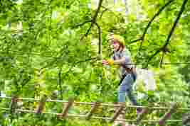 Child in a yellow helmet crossing a rope bridge in a forest adventure park, surrounded by green trees.