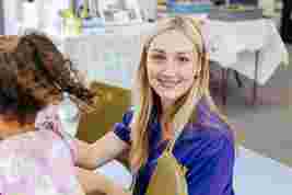 Woman in a purple shirt smiling at the camera, sitting indoors with a child facing away in the foreground. Background shows a blurred table.