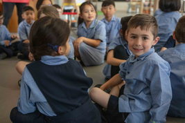 Children in school uniforms sit on the floor in a classroom, with one boy smiling at the camera amid a group of attentive classmates.
