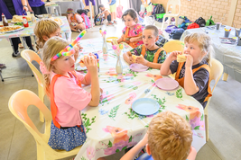 Children sit around a floral-decorated table, eating sandwiches. One child wears a colorful unicorn horn headband. Bright atmosphere.